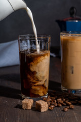Cream is poured into a glass with iced coffee, on a wooden table are coffee beans and sugar.