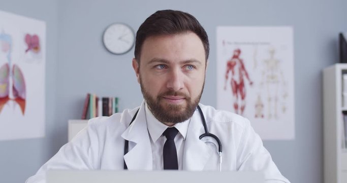 Close-up portrait young medical worker in white coat with statoscope sitting in office near computer screen. Doctor writes medical article for his blog. Сoncept of pandemic, modern medicine remotely.