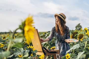 A young woman with curly hair and wearing a hat is painting in nature. A woman stands in a...