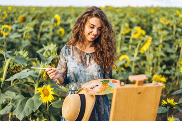 A young woman with curly hair and wearing a hat is painting in nature. A woman stands in a sunflower field on a beautiful day