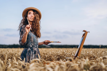 Portrait of smiling female artist with curly hair in hat. Girl draws a picture of a landscape in a...