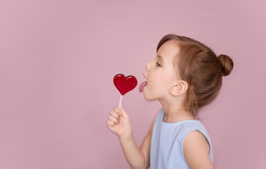 happy little girl in a blue dress licks a heart-shaped lollipop sticking out her tongue on a pink background side view