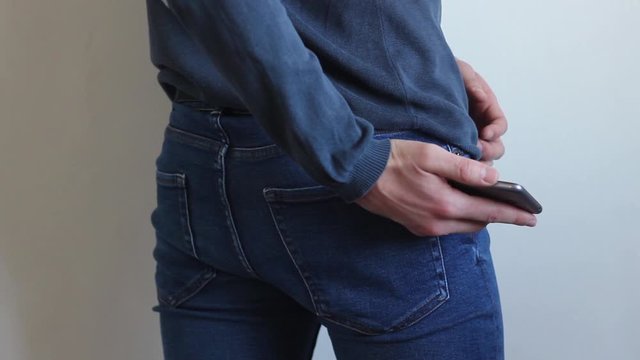 Man takes a phone from his pocket.The young man, turned back to the camera, takes his phone out of the back pocket of his jeans pants, looks at some information and puts it back in his back pocket.
