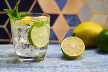 Fototapeta premium Cold refreshing summer limonade, mojito or gin tonic in glass, with fresh mint and ice cubes, lime and lemon on wooden table, on colorful background. Summer refreshment concepts.