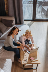 High angle view of smiling mother and kid looking at camera while using laptop near notebook on coffee table