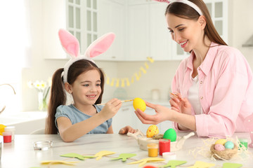 Obraz na płótnie Canvas Happy mother and daughter with bunny ears headbands painting Easter egg in kitchen