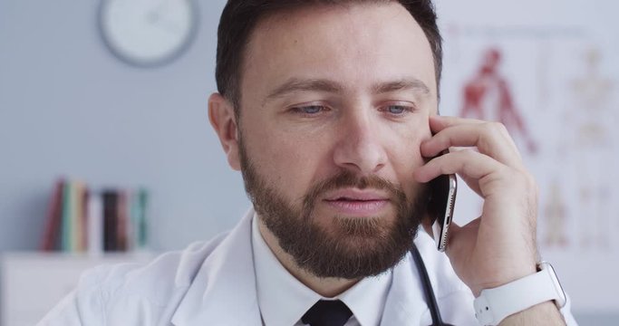 Close-up portrait of serious male doctor in lab coat and statoscope talking on mobile phone while working at desk in medical office wearing white smartwatch. Сoncept of pandemic, modern medicine.