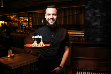 Cheerful bearded waiter with khinkali in a georgian restaurant. A discerning waiter in black clothes, with a beard and a portion of khinkali
