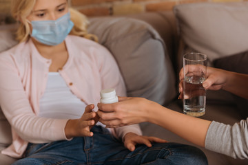Selective focus of woman giving jar with pills to daughter in medical mask on couch