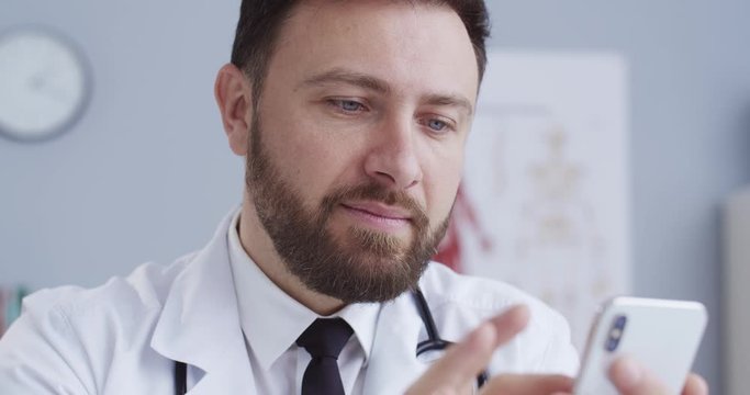 Close-up portrait of handsome doctor searching in internet. Healthcare worker reading news using smartphone. Therapist working on phone sitting in office in white medical gown with a statoscope.