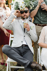 the groom in sunglasses sits on a chair in one hand holds a glass with juice from which he drinks through a straw and on whose head lies a wedding bouquet,happy wedding