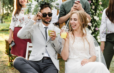 cheerful wedding couple sitting on chairs and drinking fruit drinks,wedding bouquet on the groom's head,the groom is sitting in sunglasses,cool wedding couple having fun at the wedding