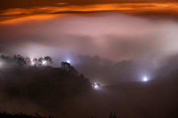 Obraz na płótnie Canvas Mountains in fog at beautiful night in autumn in Dalat city, Vietnam. Landscape with Langbiang mountain valley, low clouds, forest, colorful sky with stars, city illumination at dusk.
