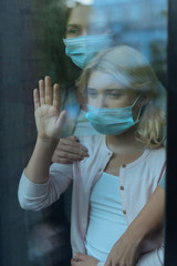 Selective focus of mother in medical mask embracing child near window at home