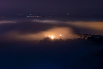 Mountains in fog at beautiful night in autumn in Dalat city, Vietnam. Landscape with Langbiang mountain valley, low clouds, forest, colorful sky with stars, city illumination at dusk.