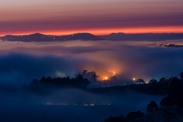 Fototapeta na wymiar Mountains in fog at beautiful night in autumn in Dalat city, Vietnam. Landscape with Langbiang mountain valley, low clouds, forest, colorful sky with stars, city illumination at dusk.