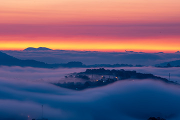 Fototapeta na wymiar Mountains in fog at beautiful night in autumn in Dalat city, Vietnam. Landscape with Langbiang mountain valley, low clouds, forest, colorful sky with stars, city illumination at dusk.