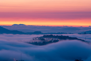 Mountains in fog at beautiful morning in autumn in Dalat city, Vietnam. Landscape with Langbiang mountain valley, low clouds, forest, colorful sky , city illumination at dusk.