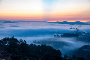 Fototapeta premium Mountains in fog at beautiful morning in autumn. Landscape with Langbiang mountain valley, low clouds, forest, colorful sky , city illumination at dusk.