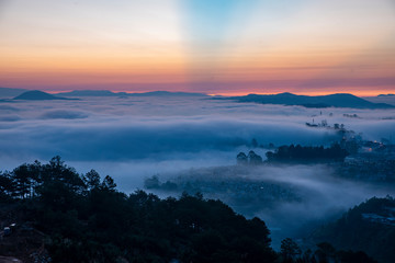 Mountains in fog at beautiful morning in autumn. Landscape with Langbiang mountain valley, low clouds, forest, colorful sky , city illumination at dusk.