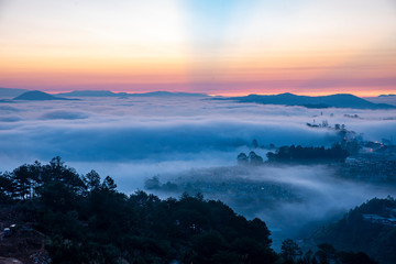 Mountains in fog at beautiful morning in autumn. Landscape with Langbiang mountain valley, low clouds, forest, colorful sky , city illumination at dusk.