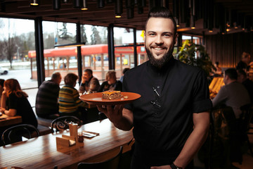 Cheerful bearded waiter with a slice of cake in a Georgian restaurant. Beautiful waiter in black clothes with a beard and a serving of cake