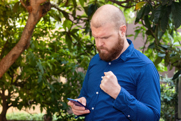 Naklejka na ściany i meble Bald bearded man in a blue shirt standing outside with an angry expression looking at a smartphone