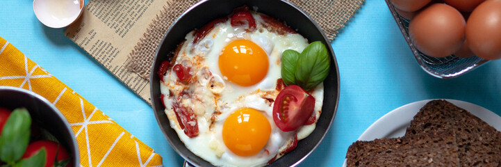 Delicious and healthy breakfast for one person: small pan with 2 fried eggs with vegetables, cheese. Served on a light bkue background with yellow napkin, fresh tomatoes and basil, brown bread. Banner