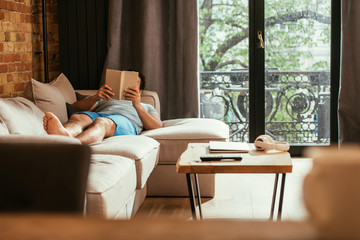 young man reading book while lying on sofa during quarantine
