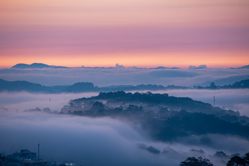 Mountains in fog at beautiful morning in autumn. Landscape with Langbiang mountain valley, low clouds, forest, colorful sky , city illumination at dusk.