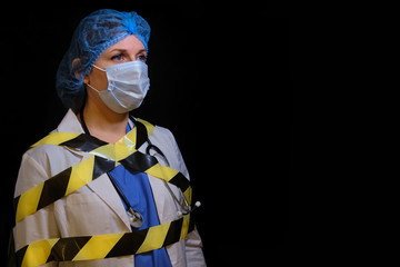 Doctor on a dark background wrapped in black and yellow stop sign tape, copy space. Portrait of a nurse in a medical mask. Concept of quarantine the coronavirus epidemic.