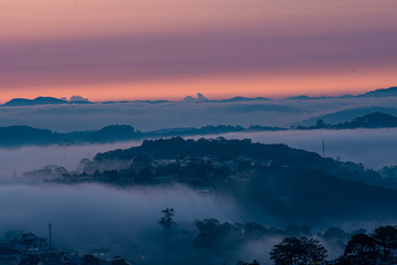 Mountains in fog at beautiful morning in autumn in Dalat city, Vietnam. Landscape with Langbiang mountain valley, low clouds, forest, colorful sky , city illumination at dusk.