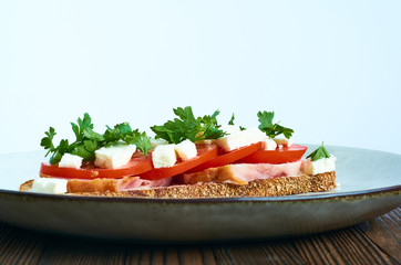 Sandwich of toast and rye bread with greens, tomatoes, feta cheese and ham in grey plate on dark wooden background.