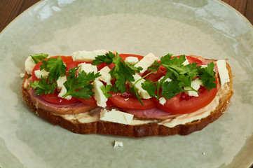 Sandwich of toast and rye bread with greens, tomatoes, feta cheese and ham in grey plate on dark wooden background.
