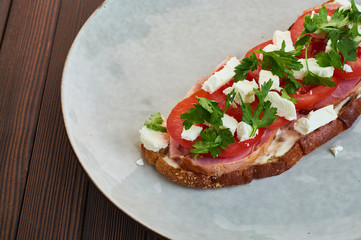 Sandwich of toast and rye bread with greens, tomatoes, feta cheese and ham in grey plate on dark wooden background.