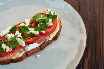 Sandwich of toast and rye bread with greens, tomatoes, feta cheese and ham in grey plate on dark wooden background.