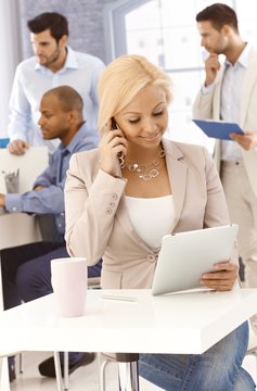 Blonde businesswoman with mobile and tablet pc in busy office.