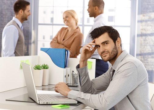 Portrait of young businessman sitting at desk, having laptop, working, looking at camera.