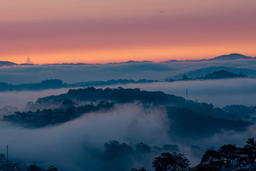 Fototapeta na wymiar Mountains in fog at beautiful morning in autumn in Dalat city, Vietnam. Landscape with Langbiang mountain valley, low clouds, forest, colorful sky , city illumination at dusk.