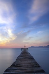 Fototapeta premium Lone person standing at end of wooden boardwalk at sunrise, with calm sea and beautiful colourful sky and mountains in the distance, Alcudia beach, Mallorca, Spain.