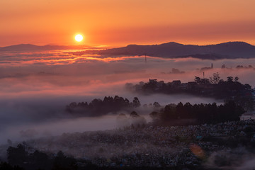 Mountains in fog at beautiful morning in autumn in Dalat city, Vietnam. Landscape with Langbiang mountain valley, low clouds, forest, colorful sky , city illumination at dusk.