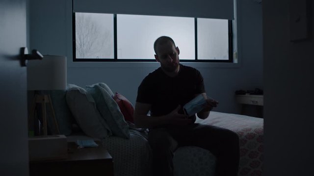 Man Sitting on Bed in Dark Room Puts Face Mask on. Wide shot Pushes In