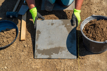 The master in yellow gloves lays paving stones in layers. Garden brick pathway paving by professional paver worker. Laying gray concrete paving slabs in house courtyard on sand foundation base.