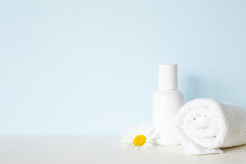 Obraz na płótnie Canvas White bottle and towel on shelf. Beautiful chamomile or daisy. Fresh flower. Care about clean and soft face, hands, legs and body skin. Empty place for text on pastel blue wall. Front view.