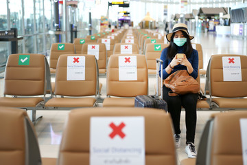 Young Asian traveler sitting at the international airport during COVID-19 disease crisis with social distancing and wearing mask. Playing mobile phone, waiting for airline counter check-in open.