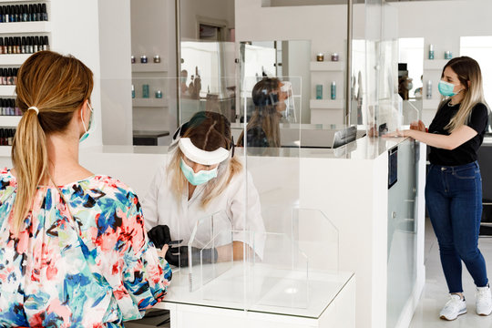 Manicure di operatrice equipaggiata con protezioni e mascherina che opera in un centro estetico attraverso una barriera divisoria di plexiglass verso una cliente bionda vestita con abito a fiori