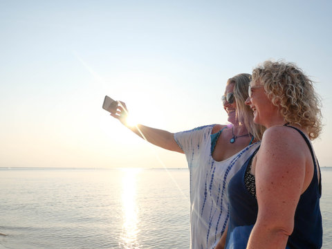 Two funny senior women making selfie on smartphone at sunset beach