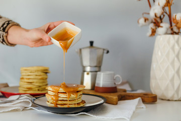 Girl pouring salted caramel on pancakes near coffee maker. Morning breakfast at home in quarantine. Recipe of homemade delicious pancakes.