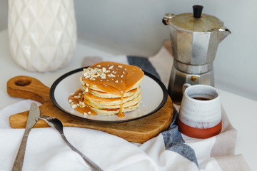 Pancakes with salted caramel and peanuts near coffee maker on white table in kitchen. Morning breakfast at home in quarantine. Recipe of homemade delicious pancakes.