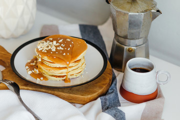 Pancakes with salted caramel and peanuts near coffee maker on white table in kitchen. Morning breakfast at home in quarantine. Recipe of homemade delicious pancakes.